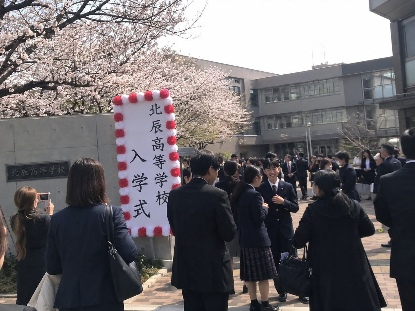 桜咲く校門の入学式看板の前で記念撮影する新入生